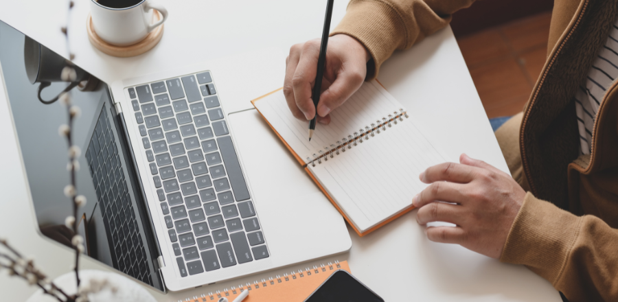 Laptop open with person taking notes in notebook in front  