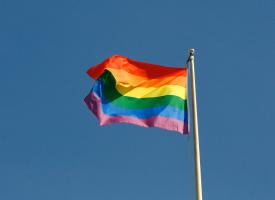 A rainbow pride flag on a flagpole waves in front of a blue sky