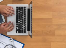 Computer open, birds eye view with hands typing and stethoscope to one side. 