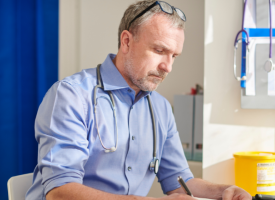 Image of a male doctor at a desk in a GP practice writing in a notebook