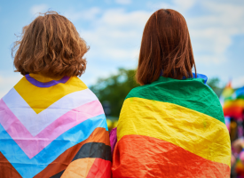 Two people with their backs to the camera, wearing LGBTQIASB+ flags over their shoulders