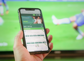 Phone with a gambling app open in front of TV showing sport
