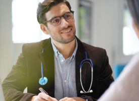 A male doctor with glasses wearing a stethoscope holds a pen over a notepad, listening to a woman.