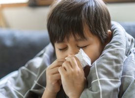 A young boy sneezes into a tissue, wrapped in a blanket.