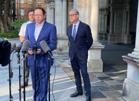 Professor Paul Griffin stands in front of microphone stands. Health Minister Tim Nicholls stands to his left.