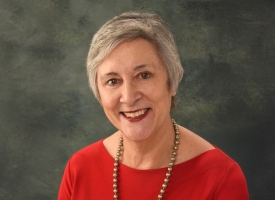A woman with short grey hair and a broad smile looks at the camera. She is wearing a red blouse and gold necklace.