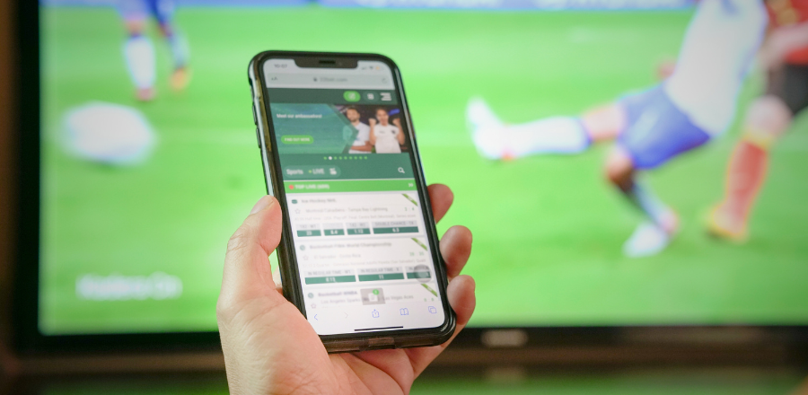 Phone with a gambling app open in front of TV showing sport